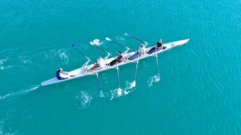 Blick von oben auf 5 Personen in einem Ruderboot auf dem Wasser 4 Personen rudern