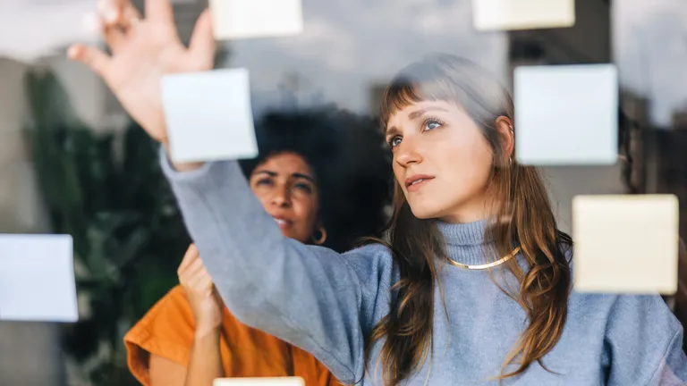 Two women in a modern office collaborating at a glass wall covered with sticky notes while planning a project.